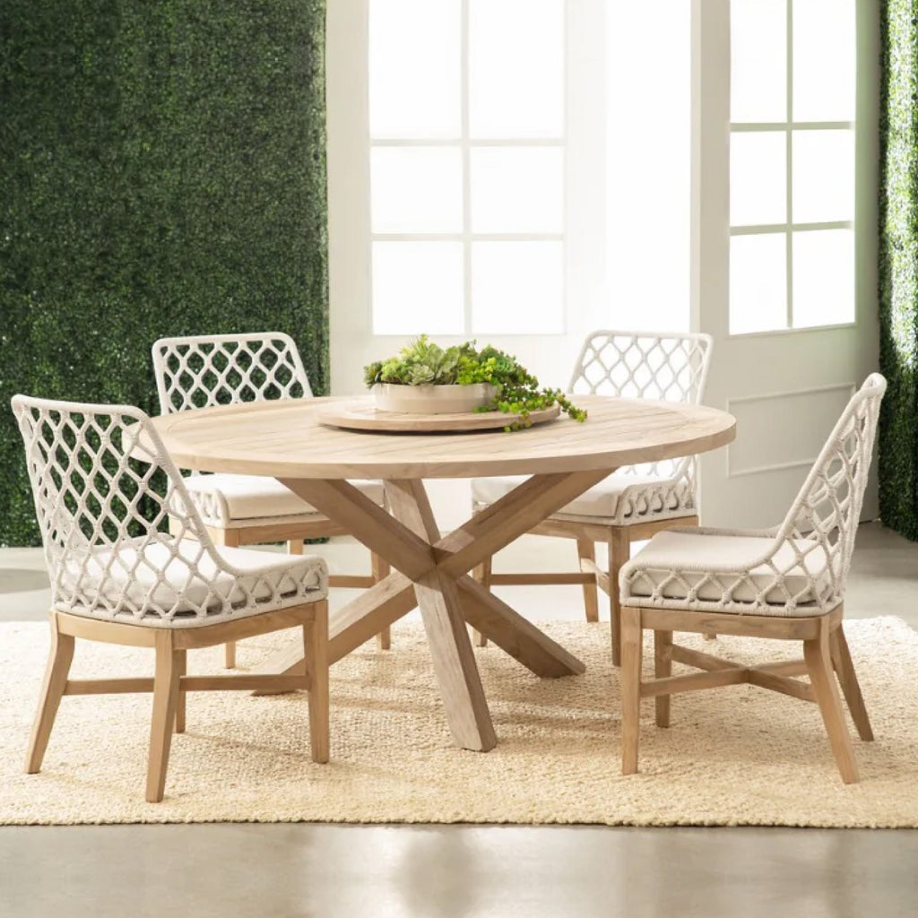 A round wooden dining table with four lattice-back chairs, set on a beige rug, against a green hedge wall.