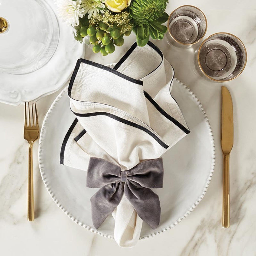 Elegant dining setup with a white plate, gold cutlery, and a folded napkin with a gray bow, garnished with flowers.