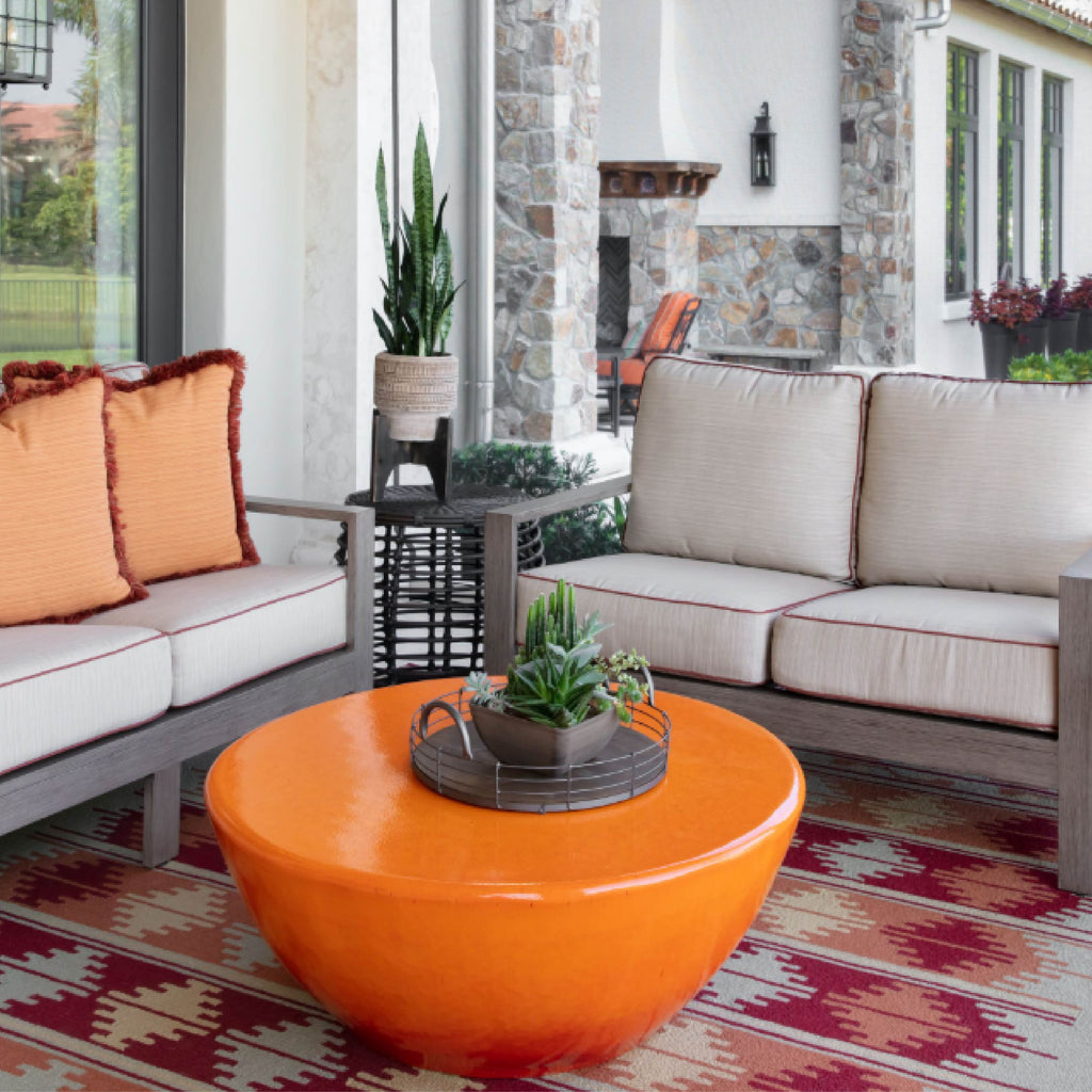 Outdoor seating area with two sofas, an orange round table, and potted plants on a patterned rug.