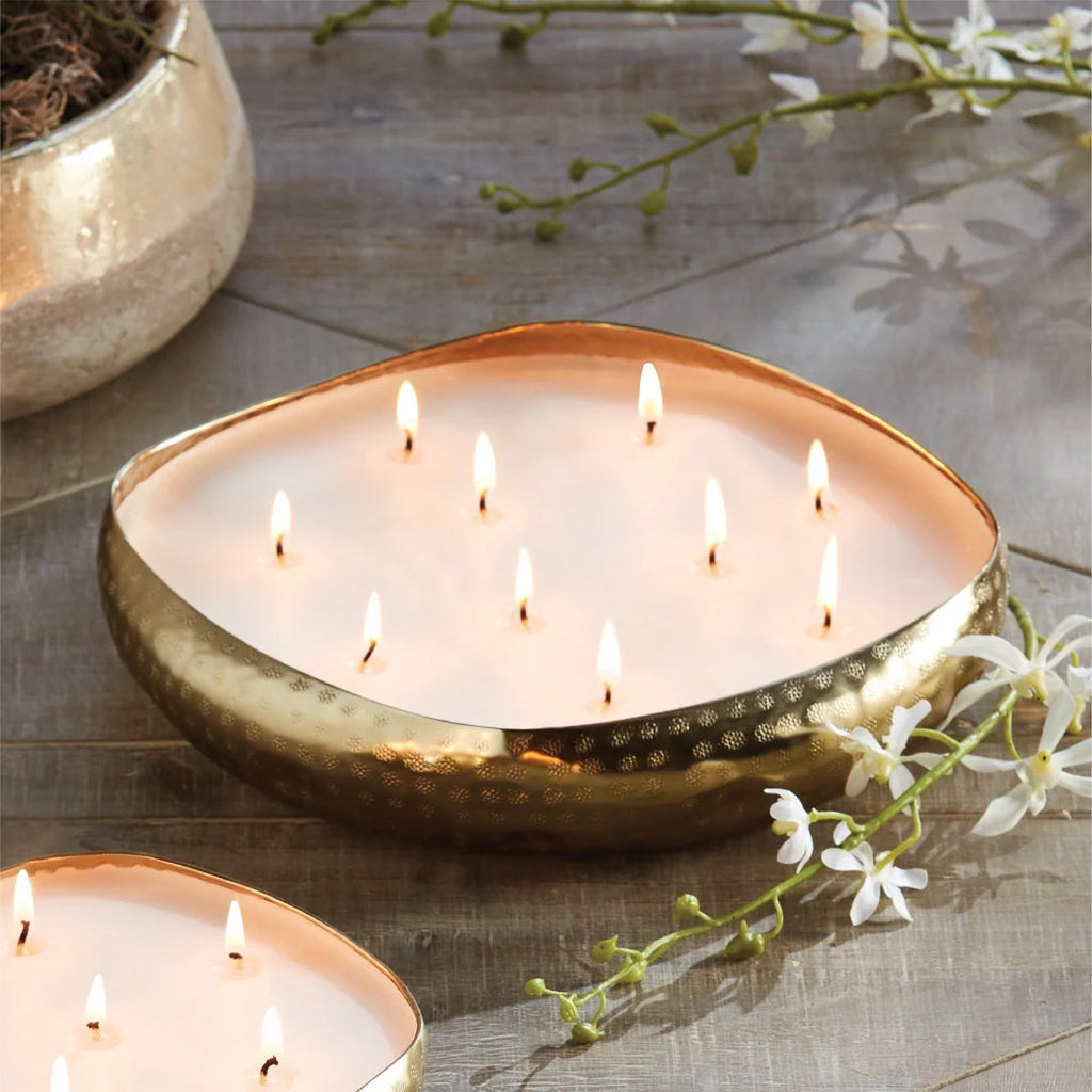 Oval-shaped candle with multiple flames in a gold holder, surrounded by white flowers on a wooden surface.