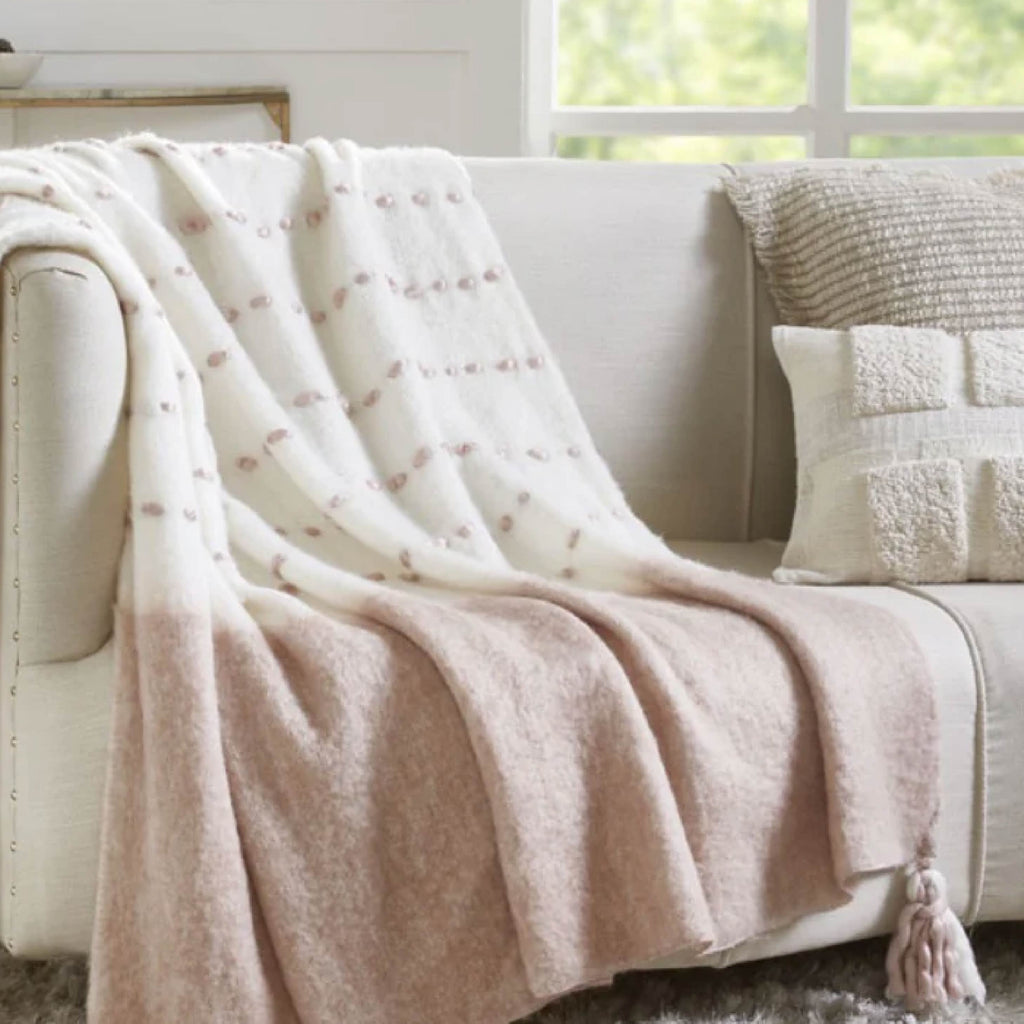 A beige sofa with a pink and white blanket and a textured pillow, near a window.