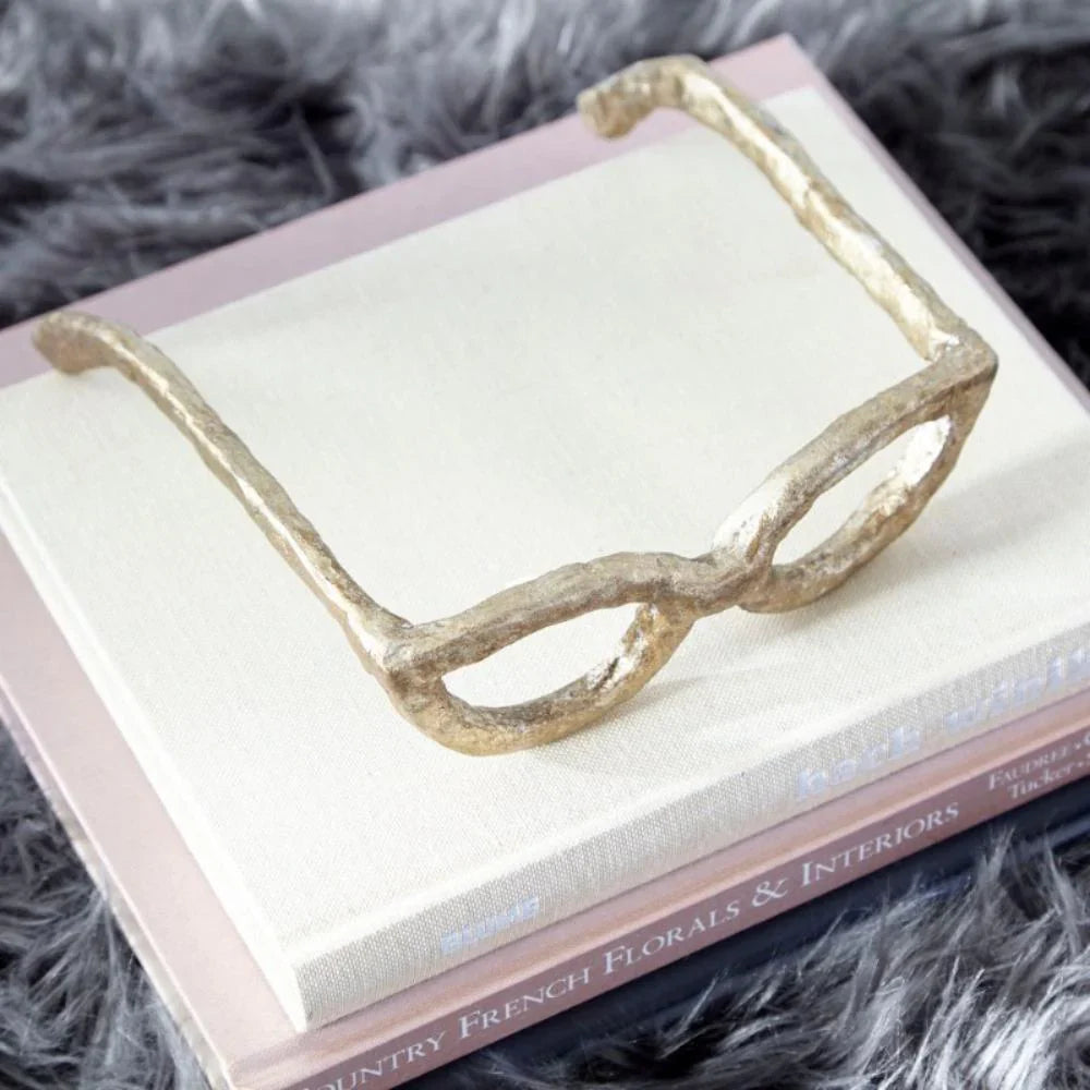 Bronze sculpture of glasses on a stacked two books, one with a white cover and the other pink, on a gray fuzzy surface.