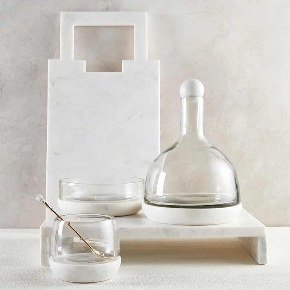 Marble cutting board, glass decanter, and bowls with a decorative spoon on a white surfaced background.