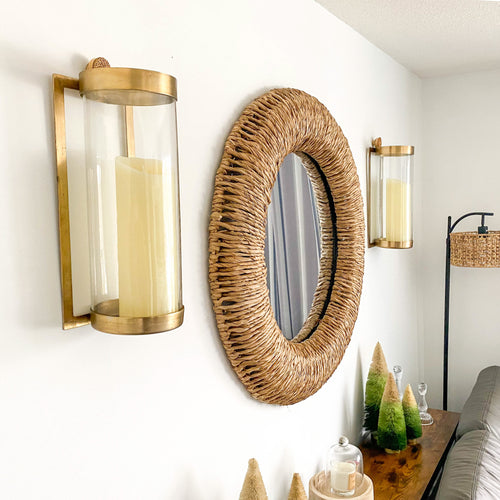 A living room wall is beautifully adorned with a woven mirror in antique brass, decorative trees, and Kalalou's Glass + Antique Brass Candle Wall Sconces, complemented by a wall-mounted candle lantern.