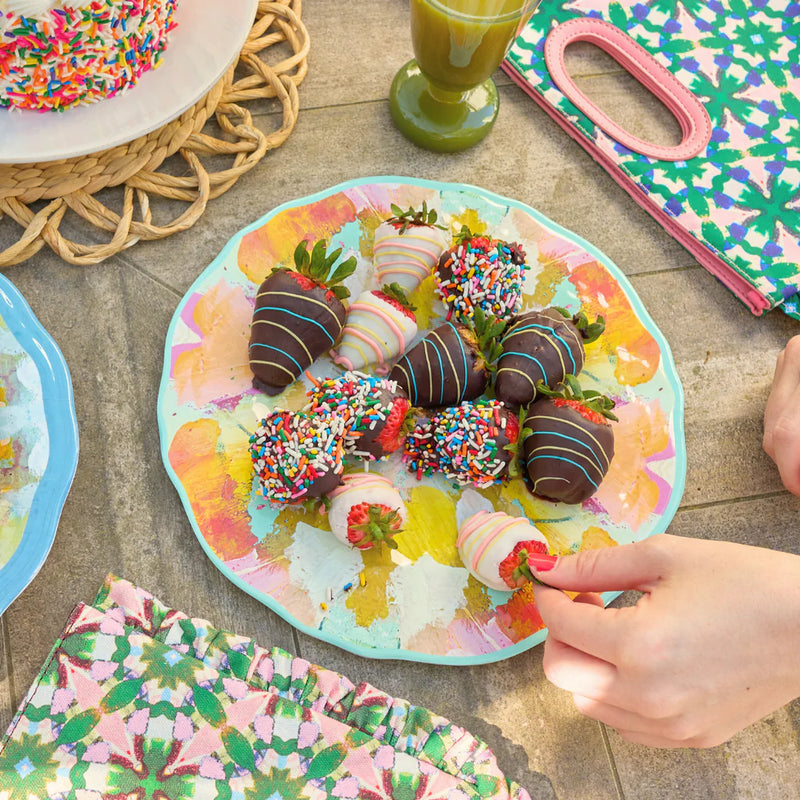 A hand reaches for a colorful chocolate-covered strawberry with sprinkles, artfully arranged on a Marigold Melamine Pasta Bowl by Laura Park Designs. Perfect for vibrant and BPA-free table settings.