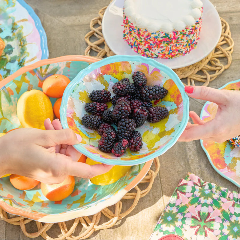 Hands exchanging a colorful Marigold Melamine Pasta Bowl filled with blackberries; fruits and a sprinkle-covered cake add charm to the vibrant table setting by Laura Park Designs.