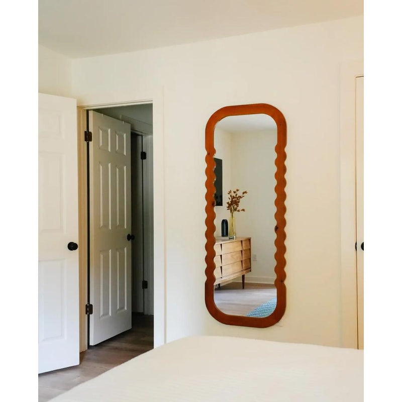 A minimal bedroom scene featuring the Kalalou Natural Wood Scalloped Floor Mirror, an open door, and a dresser adorned with a vase of dried flowers.