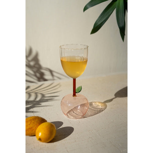 An Apple Base Wine Glass from Kalalou, with orange liquid and a leaf, sits on a table next to two lemons casting leaf shadows.