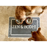 Two dogs eating from bowls on a Spicher and Company Mosaic 46th Street Personalized Vinyl Mat, featuring a vintage black and white tile design with white text reading "ZEN & BODHI." The mat is available in multiple sizes, easy to clean, and adds a touch of style to your pet's dining area.