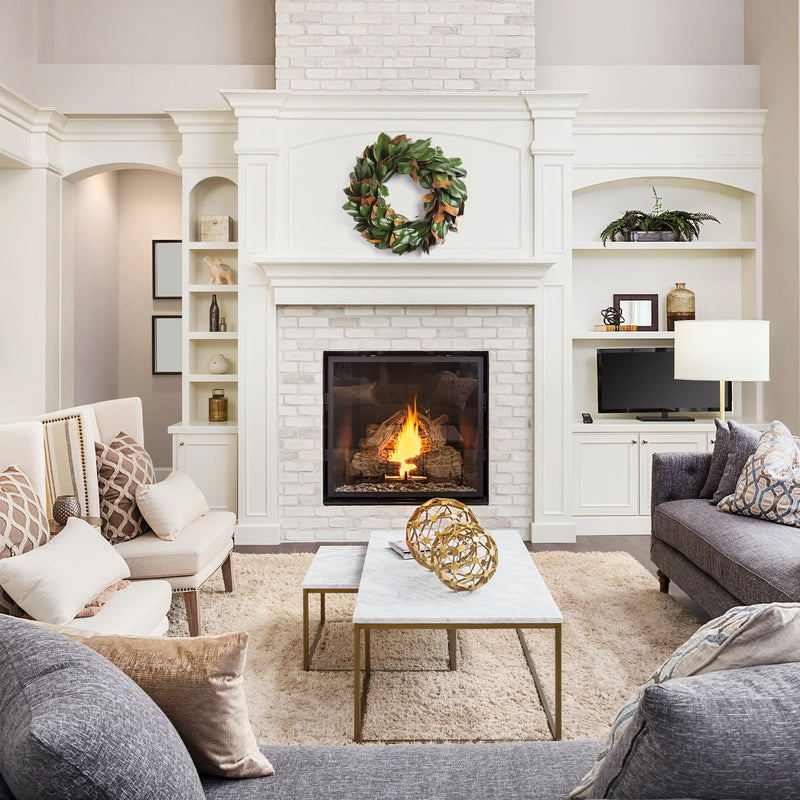 A cozy living room with a fireplace, white brick mantel, and modern decor. Neutral-colored furniture and the Grand Magnolia Leaf Wreath by Napa Home & Garden above add an elegant touch for interior use.