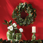 A Grand Magnolia Leaf Wreath by Napa Home & Garden adorns a red wall, complemented by green vases holding white flowers and a lit white candle on the table, creating a charming scene perfect for interior use.