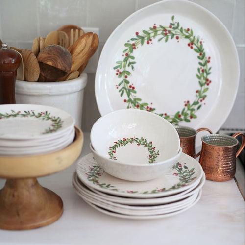 White ceramic dishes adorned with green leaves and red berries arranged on a countertop near mugs and wooden utensils create a festive display, complemented by the Always Relish Christmas Wreath Melamine Serving Platters (set of 2).